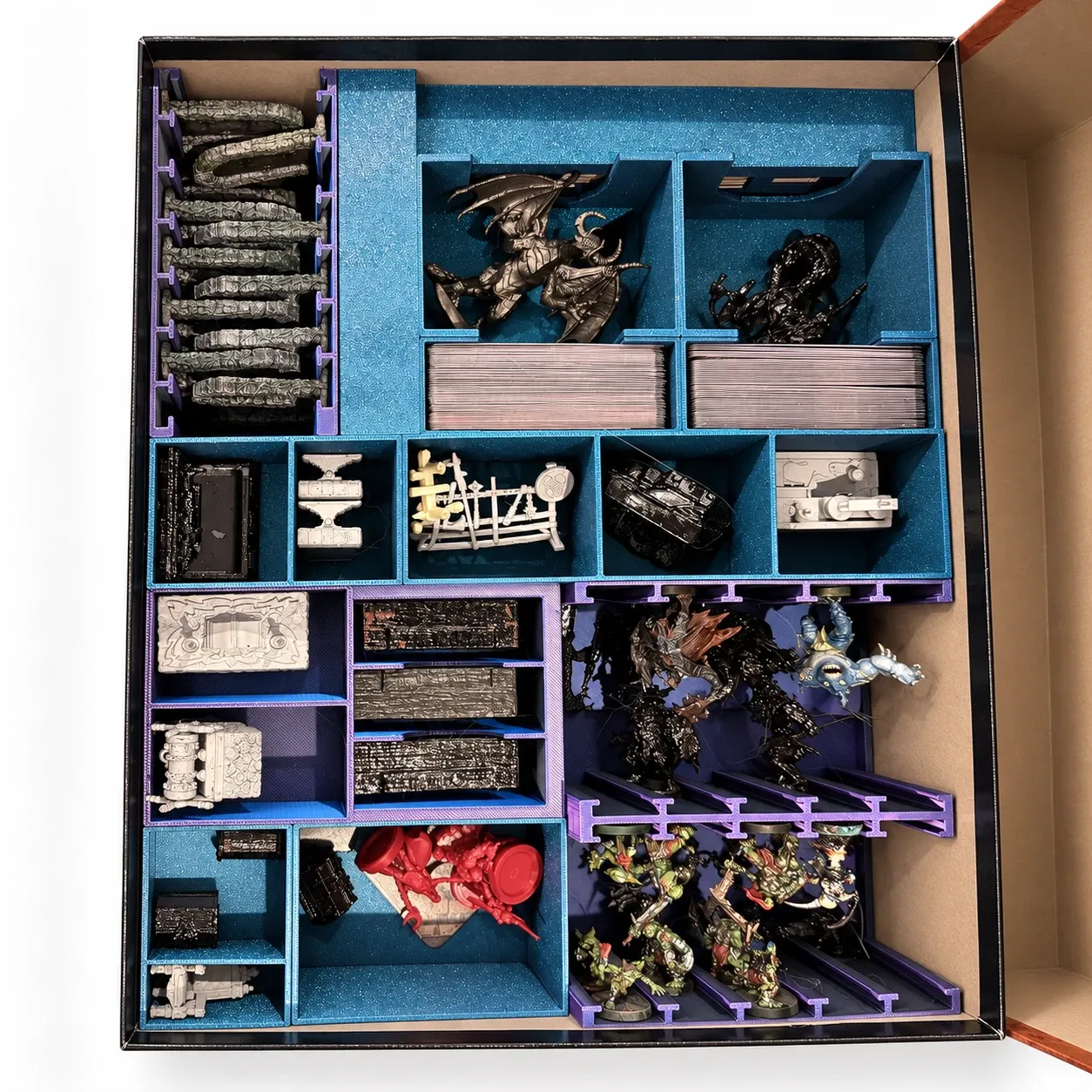 Inserto organizador impreso en 3D dentro de la caja de un juego de mesa, con compartimentos azules y morados para miniaturas, cartas y losetas
