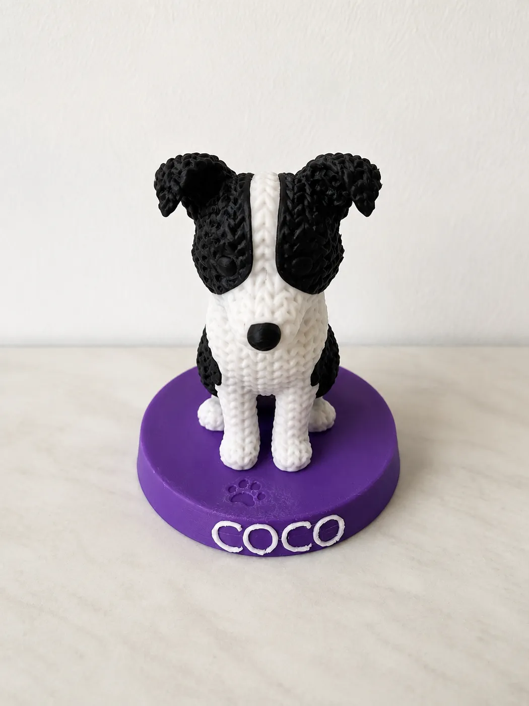 Figura de border collie con textura de crochet sobre peana morada con el nombre Coco, impresa en 3D